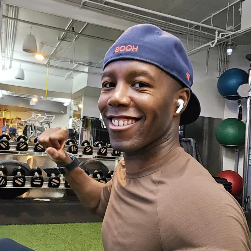 Pierre smiling in a gym, wearing a brown T-shirt and a backward baseball cap.